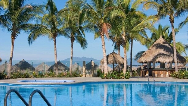 A swimming pool with clear blue water, surrounded by palm trees and thatched-roof structures. - CONDOMINIO GRAND MARINA by TRVL2HM (Nuevo Nayarit)