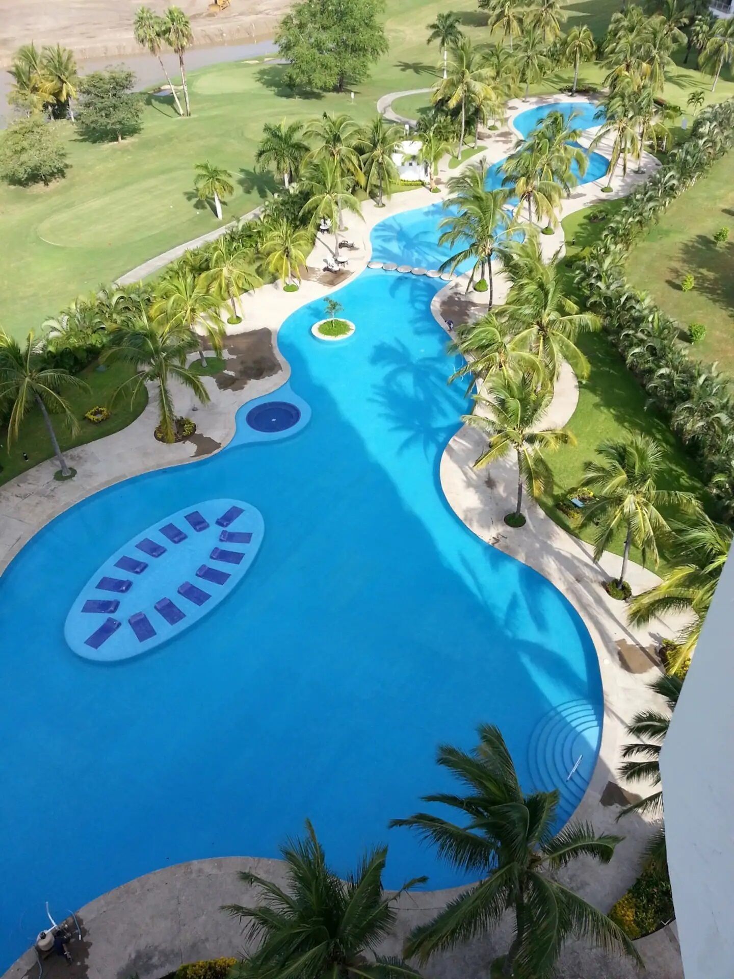 Une grande piscine sinueuse, bordée de palmiers, avec un terrain de golf en arrière-plan.