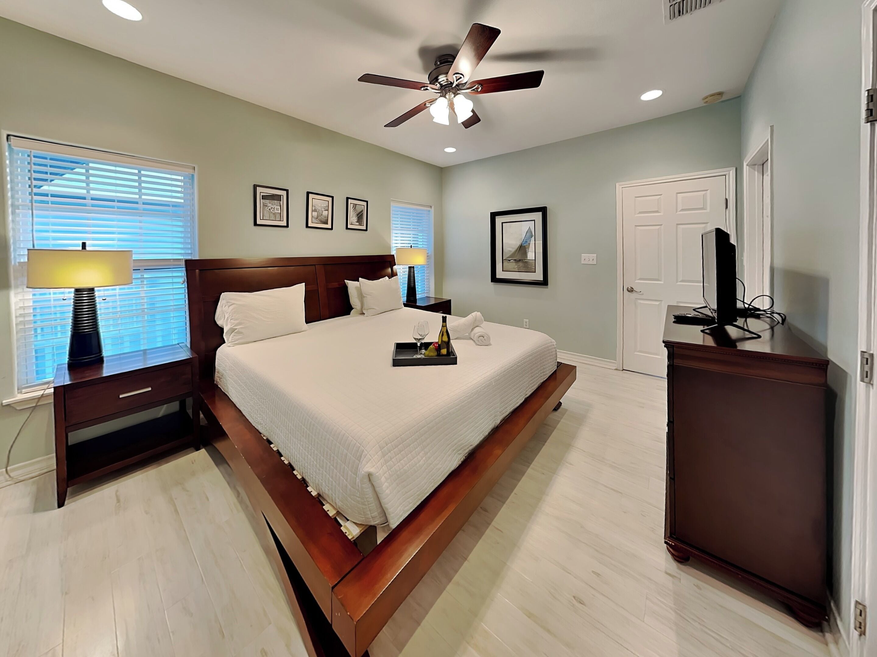 A hotel room with a bed, bedside lamps, a ceiling fan, a television, and framed pictures on the wall.