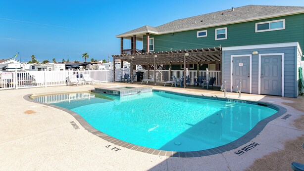 A swimming pool with a wooden deck and lounge chairs, surrounded by a fenced area with a building in the background. - Beach Bunker by Silver Sands Vacation Rentals (Port Aransas)