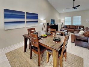 A dining area with a wooden table set for four, surrounded by chairs. There are two wall-mounted paintings, a ceiling fan, and a window with blinds. - Beach Bunker by Silver Sands Vacation Rentals (Port Aransas)