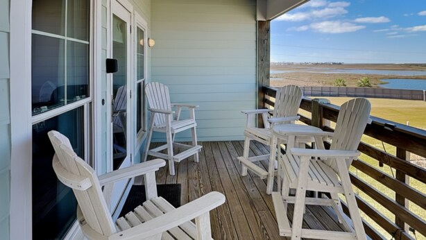 Un porche cubierto con sillas Adirondack blancas, una terraza de madera y vistas a una playa y un humedal.