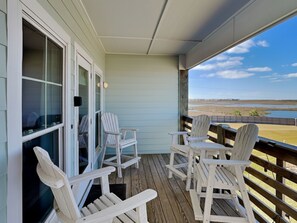 Eine überdachte Veranda mit weißen Adirondack-Stühlen, einer Holzterrasse und Blick auf Strand und Marschland.