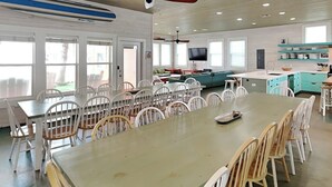 A spacious dining area with a long table and multiple chairs, a kitchen area with teal cabinets, and a television mounted on the wall. - Beach Club 2 by Silver Sands Vacation Rentals (Port Aransas)
