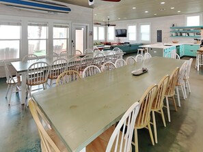 A spacious dining area with a long table and multiple chairs, a kitchen area with teal cabinets, and a television mounted on the wall. - Beach Club 2 by Silver Sands Vacation Rentals (Port Aransas)