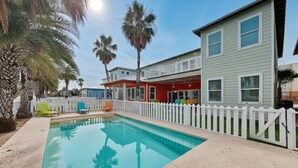 A swimming pool with a white picket fence, surrounded by palm trees and houses with a red and grey facade. - Beach Club 2 by Silver Sands Vacation Rentals (Port Aransas)