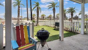 A covered patio with a grill, a colorful bench, and a view of a fenced yard with palm trees. - Beach Club 2 by Silver Sands Vacation Rentals (Port Aransas)