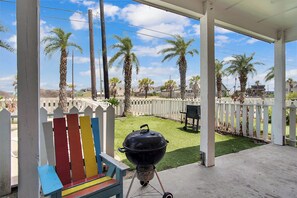 A covered patio with a grill, a colorful bench, and a view of a fenced yard with palm trees. - Beach Club 2 by Silver Sands Vacation Rentals (Port Aransas)