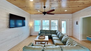 A modern living room with a sectional sofa, a flat-screen TV, a ceiling fan, and large windows. - Beach Club 1 by Silver Sands Vacation Rentals (Port Aransas)