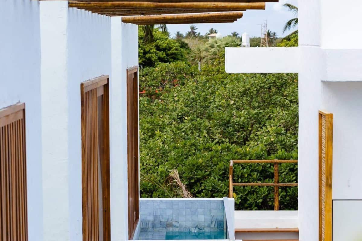 A modern outdoor pool area with a wooden pergola, white walls, and a lush green backdrop.