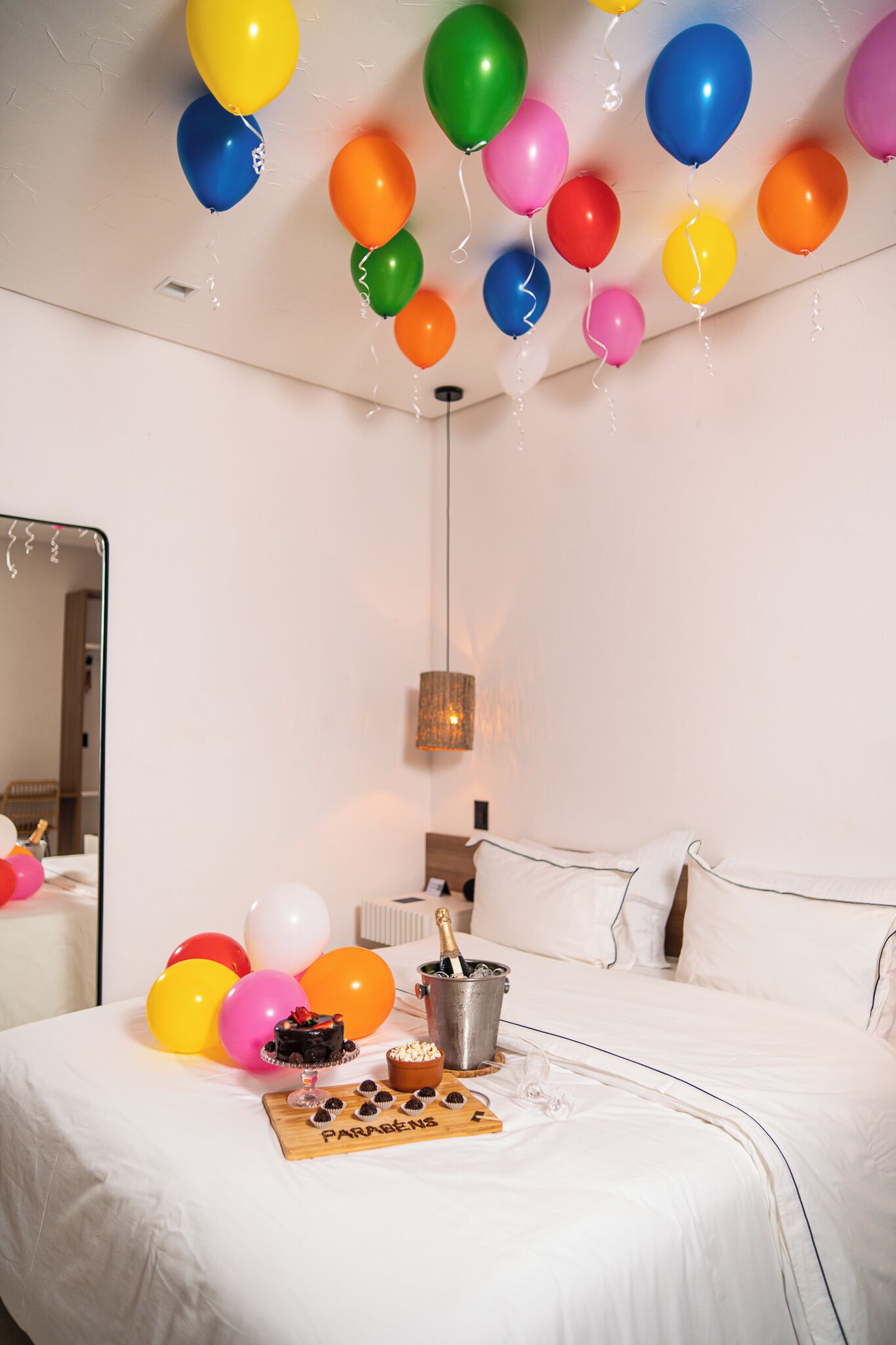 A hotel room with a bed, a bedside table with a champagne bottle, and colorful balloons on the ceiling.