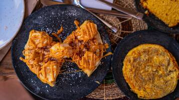 Un plato negro con un alimento cubierto de salsa, un plato pequeño con un guiso al horno, y una cuchara sobre un tapete tejido.