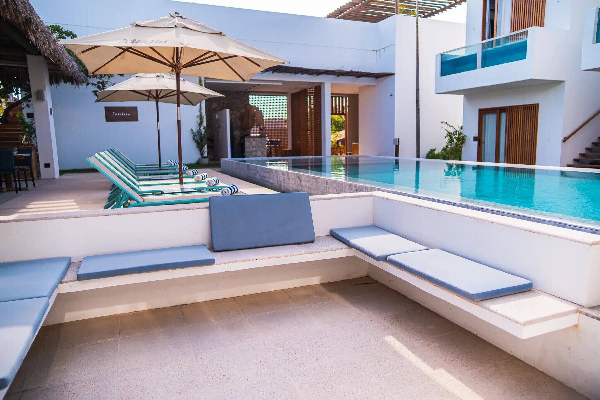 Un área de piscina moderna con sillones de descanso y sombrillas, un muro de piedra y una escalera que conduce a un segundo piso.