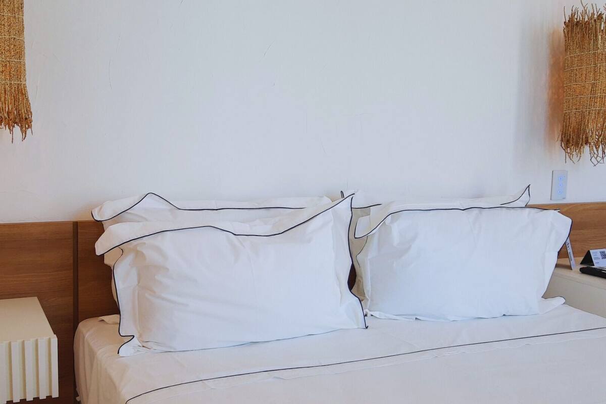 A neatly made bed with white bedding and black piping, a bedside table with a remote control, and a wall-mounted light fixture.