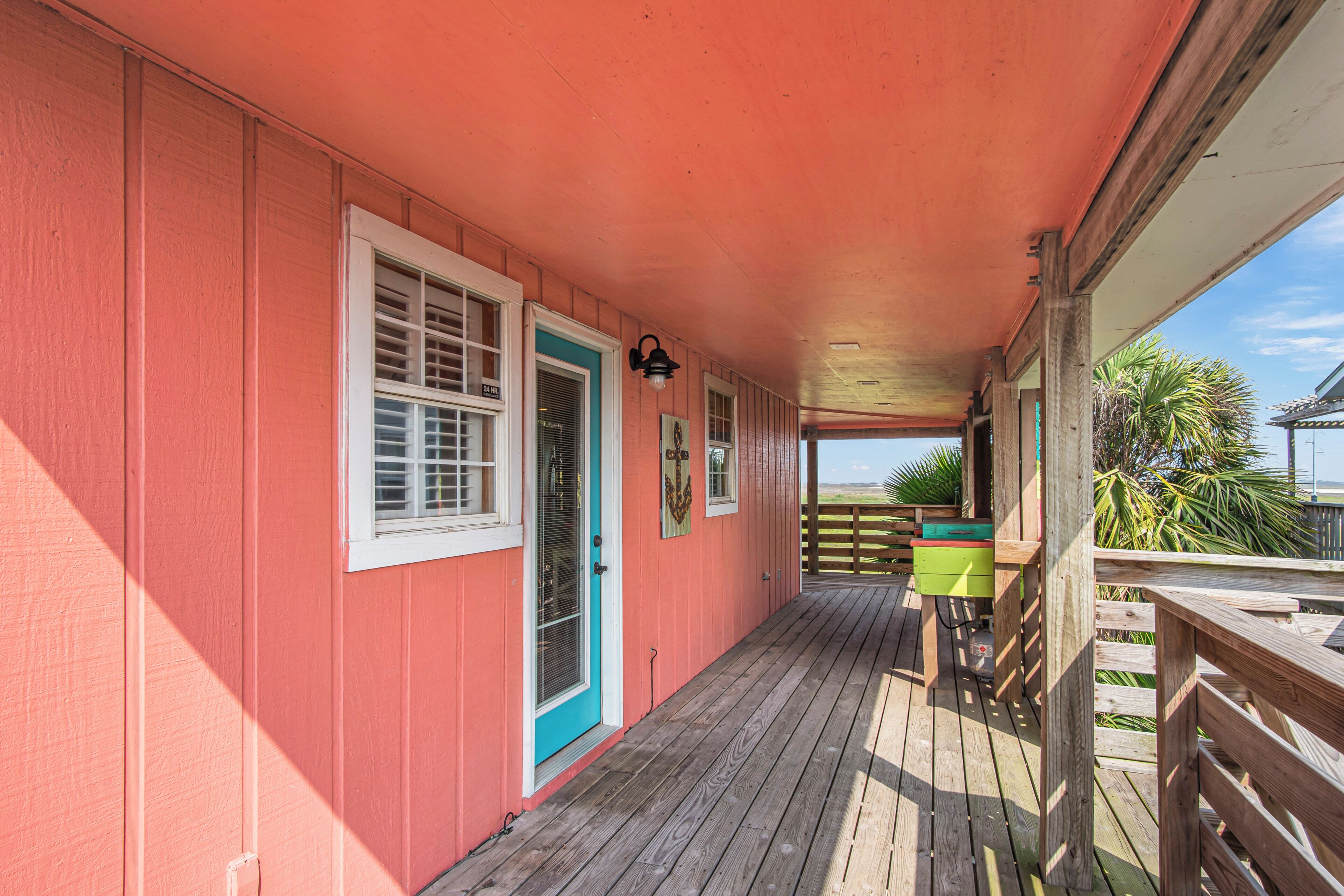 Une terrasse en bois couverte, avec un mur extérieur rouge, une porte dotée d’un cadre bleu et une poubelle verte.