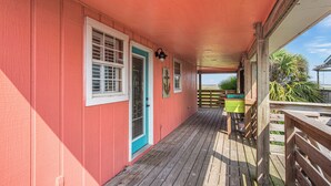 A covered wooden deck with a red exterior wall, a door with a blue frame, and a green trash bin. - Beachin' Country by Silver Sands Vacation Rentals (Port Aransas)