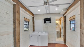 A laundry room with two white washers and dryers, wooden beams, a ceiling fan, and a TV mounted on the wall. - Beachin' Country by Silver Sands Vacation Rentals (Port Aransas)