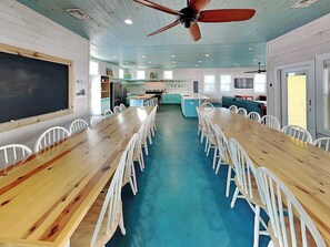 A long wooden table with white chairs, a blackboard, and a kitchen area with teal cabinets.