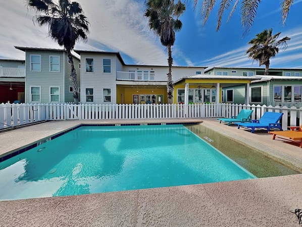 A swimming pool with a white fence, blue lounge chairs, and palm trees.