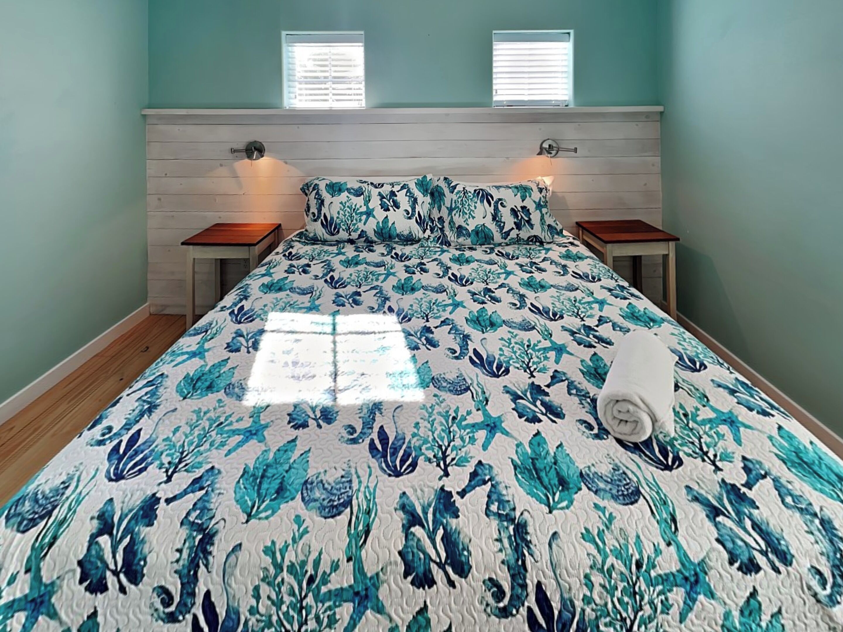A bedroom with a bed, two nightstands, a wooden headboard, and two windows with blinds.