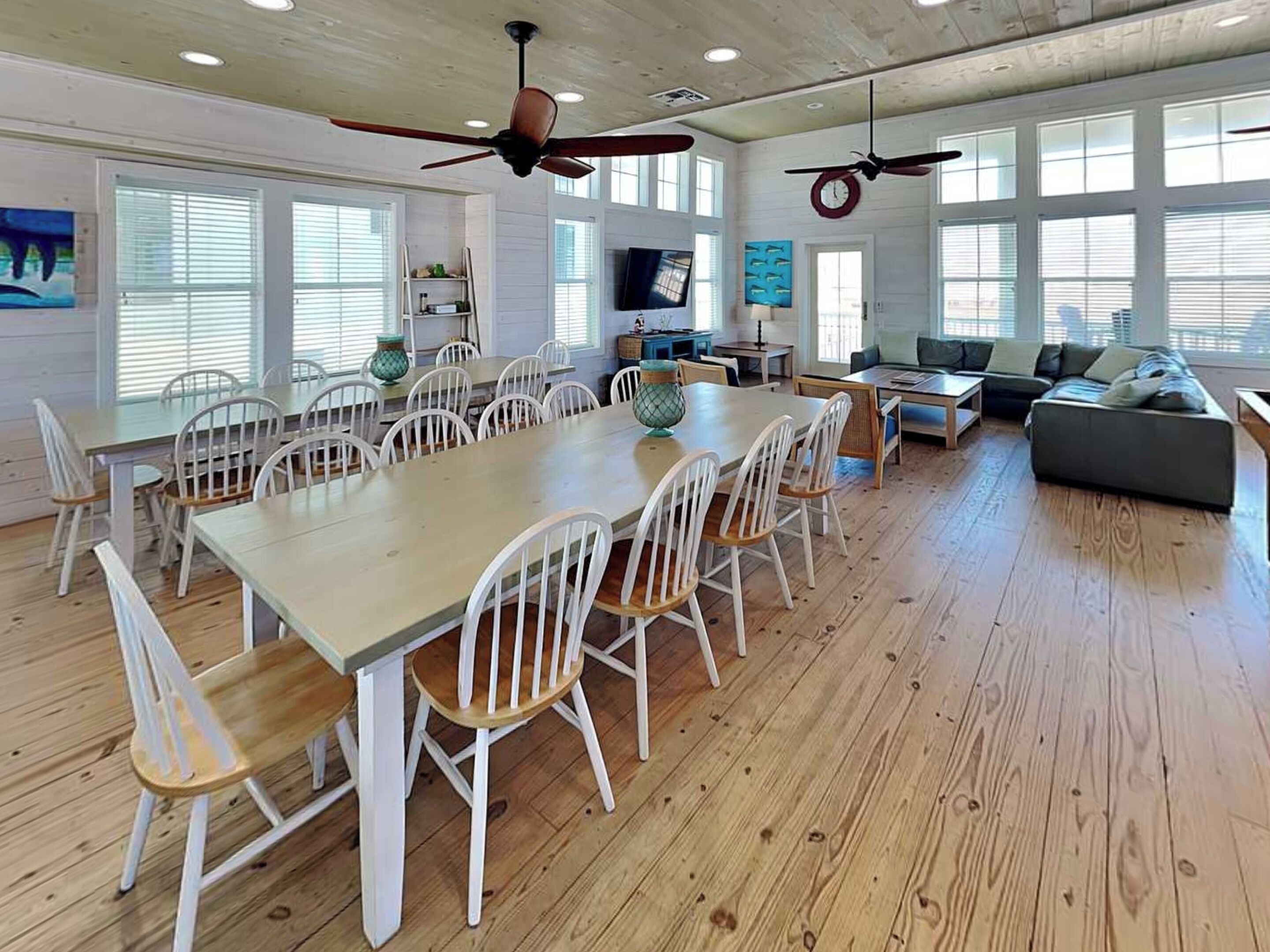 A spacious dining area with a long table and white chairs, a living area with a sofa and a television, and a ceiling fan.