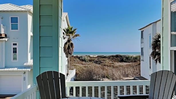 A wooden deck with a round table and two chairs, overlooking a beach with houses in the background. - Bella Vista by Silver Sands Vacation Rentals (Port Aransas)