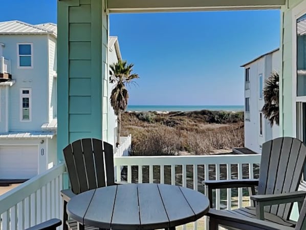 Um deck de madeira com uma mesa redonda e duas cadeiras, com vista para uma praia e casas ao fundo.