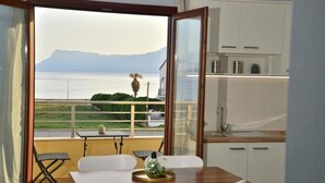 A balcony with a view of the sea and mountains, a dining area with a table and chairs, and a kitchen with white cabinets.