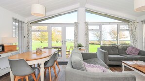 Un salon spacieux avec une grande table à manger, des canapés gris et une vue sur un paysage verdoyant grâce à de grandes fenêtres.