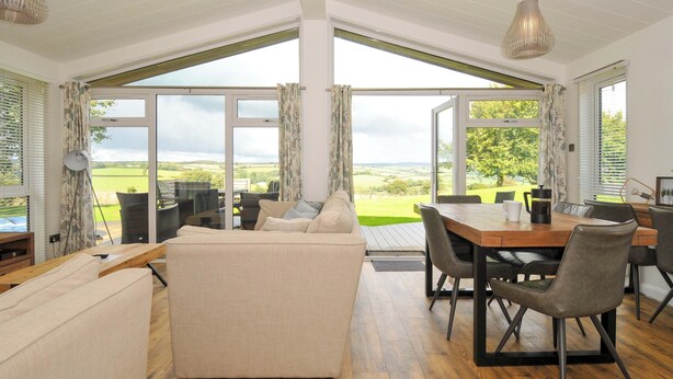 A spacious living area with a large sofa, dining table, and chairs, featuring large windows with a countryside view. - 8 Horizon View (Liskeard)
