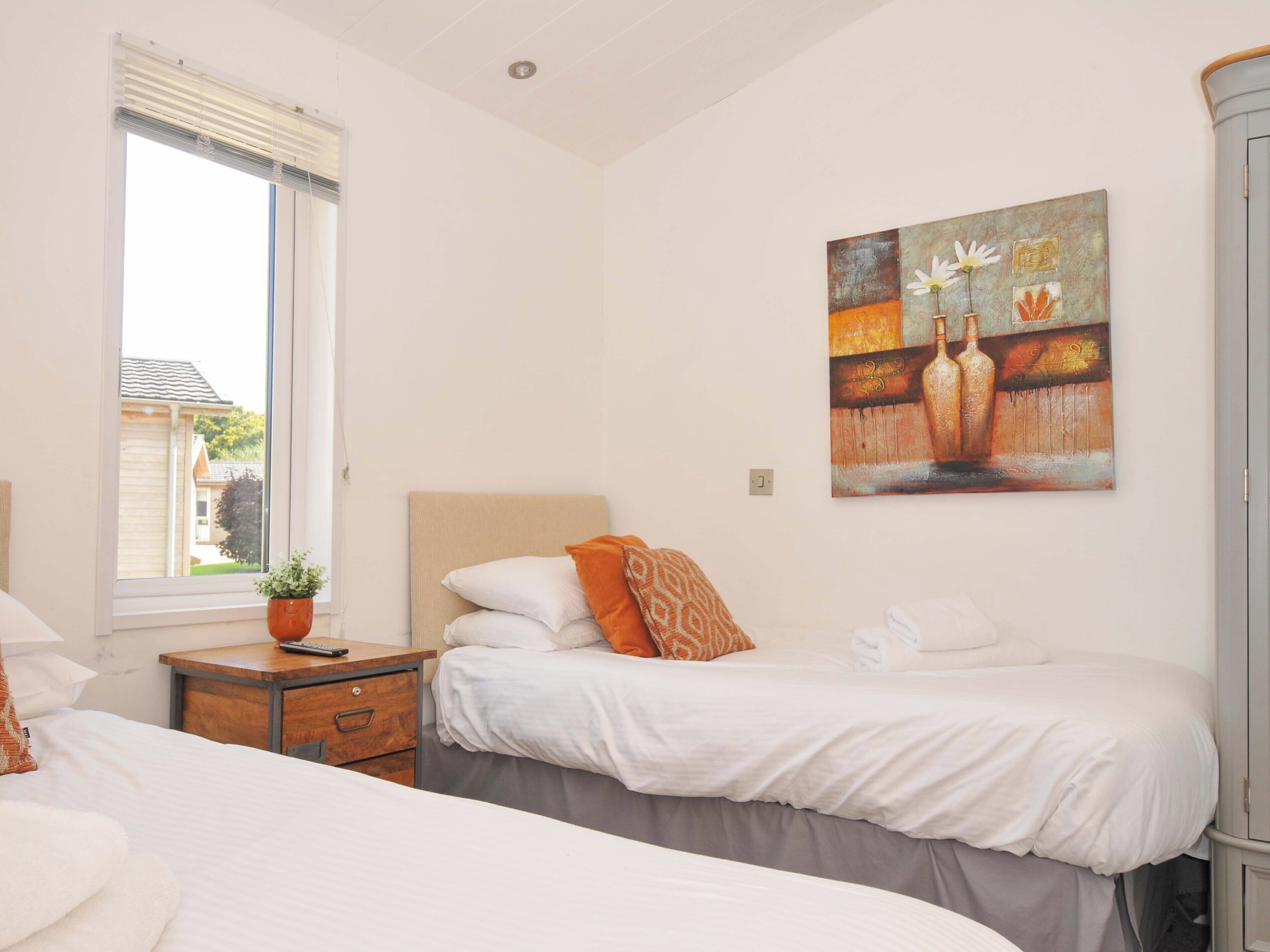 A bedroom with two beds, a window with blinds, a bedside table with a plant, and a painting on the wall.