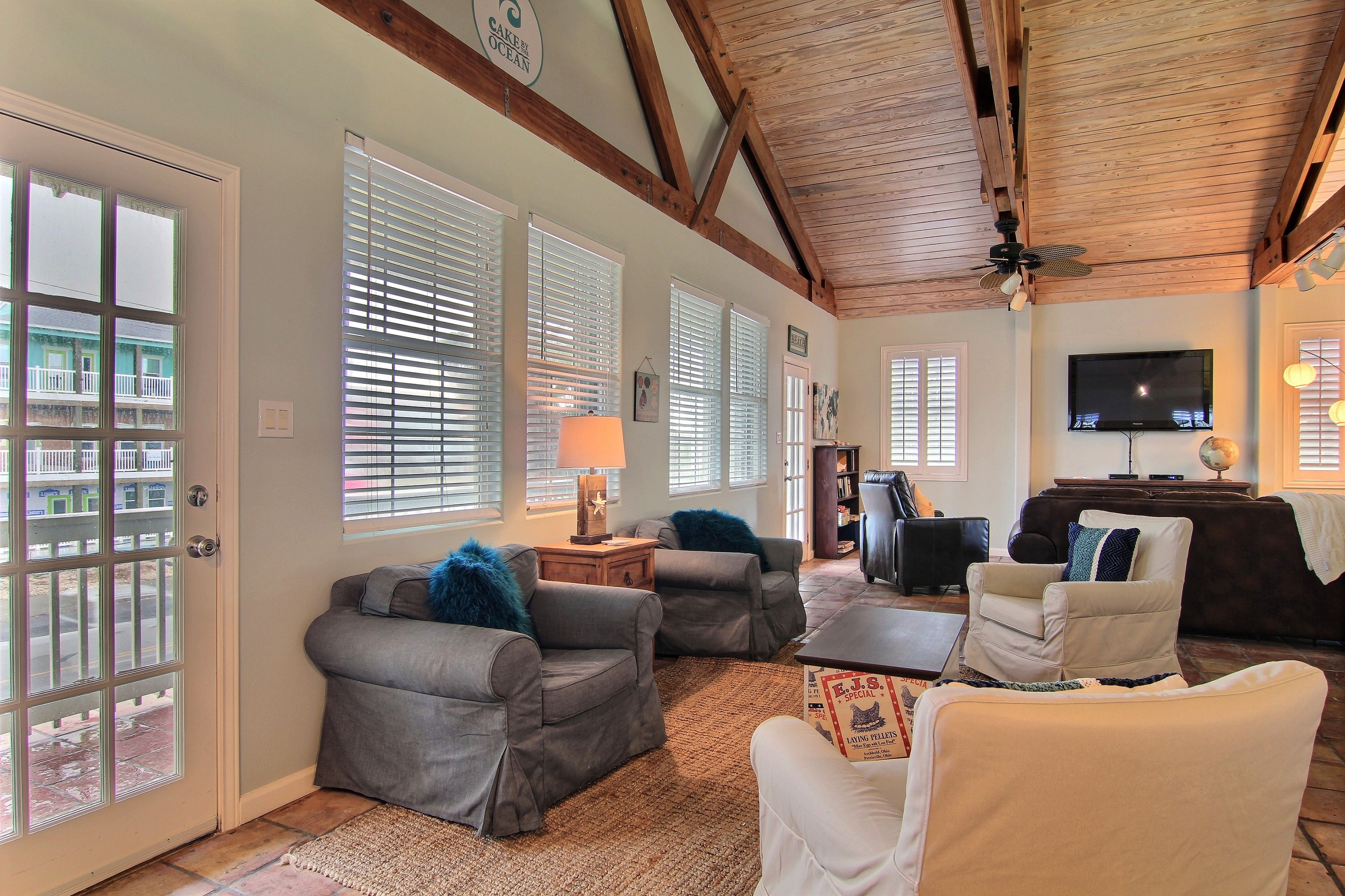 A spacious living room with a high ceiling, large windows with blinds, and a flat-screen TV mounted on the wall.