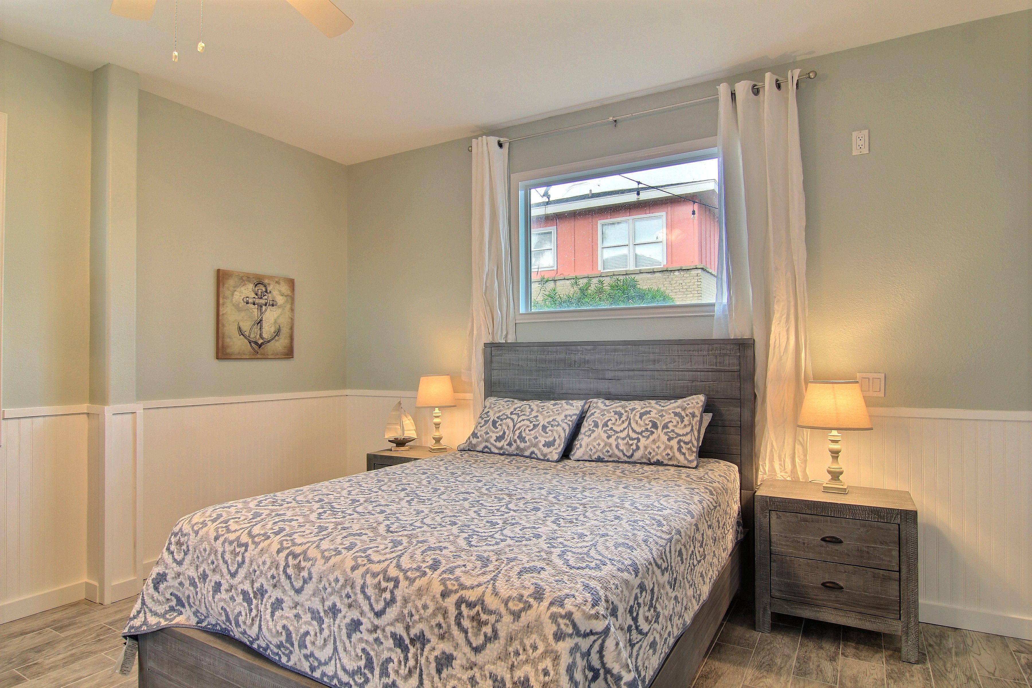 A bedroom with a bed, two bedside tables with lamps, a window with curtains, and a wall-mounted artwork.