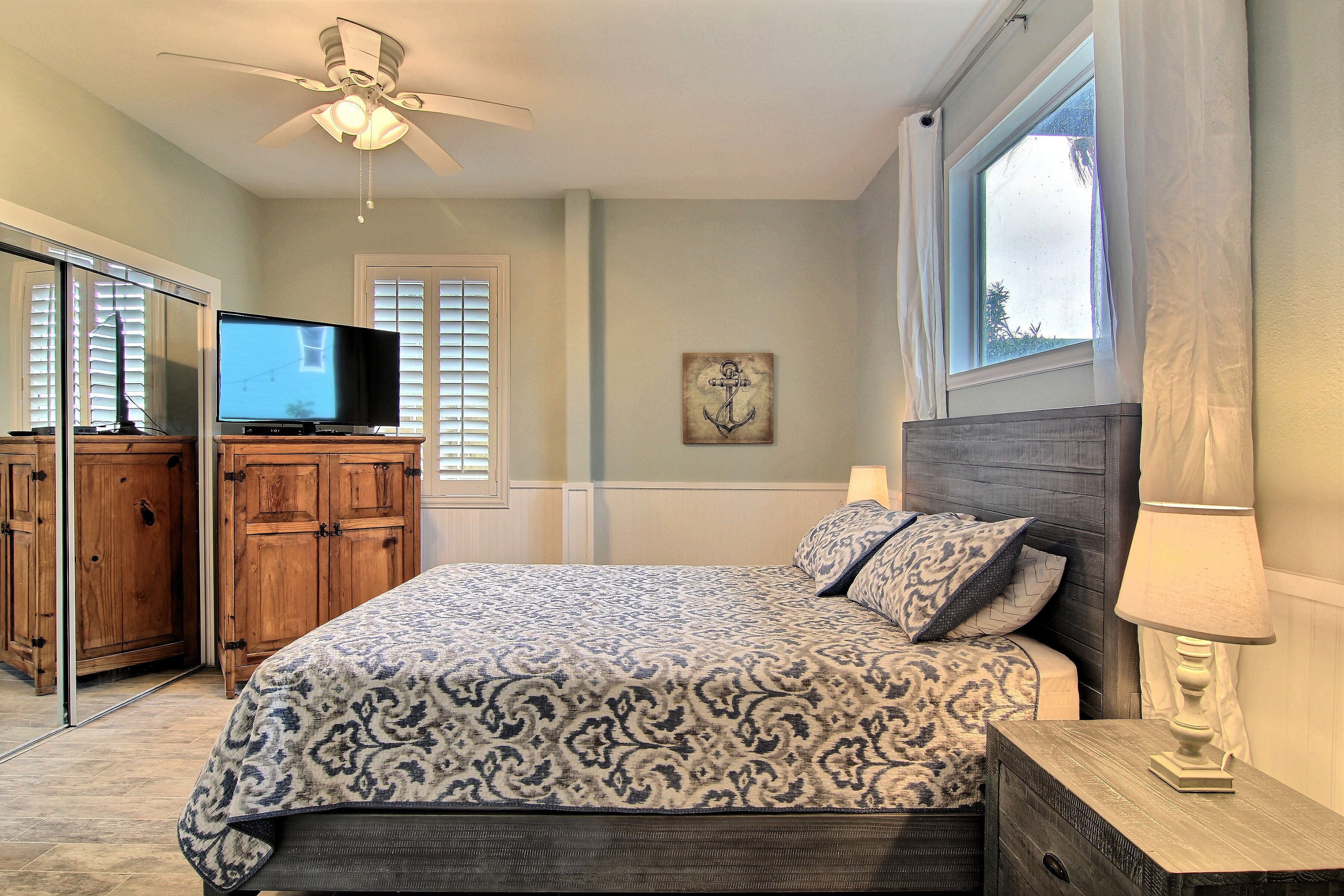 A bedroom with a bed, a television, wooden cabinets, a ceiling fan, and a window with curtains.