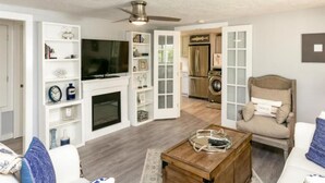 A modern living room with a fireplace, television, and a dining area with a kitchen in the background. - Cape Cod Cottage by Silver Sands Vacation Rentals (Port Aransas)