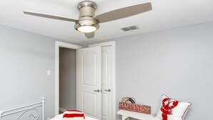 A bedroom with a bed, a ceiling fan, a door, and a small table with a sign. - Cape Cod Cottage by Silver Sands Vacation Rentals (Port Aransas)