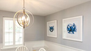 A dining area with a white table, white chairs, a chandelier, and two framed artworks on the wall. - Cape Cod Cottage by Silver Sands Vacation Rentals (Port Aransas)