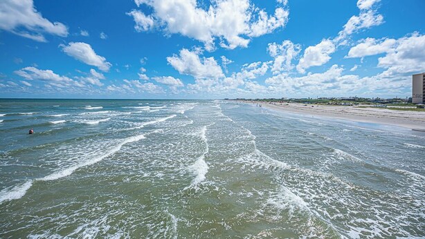 A beach with waves, a clear sky, and distant buildings. - Cape Cod Cottage by Silver Sands Vacation Rentals (Port Aransas)