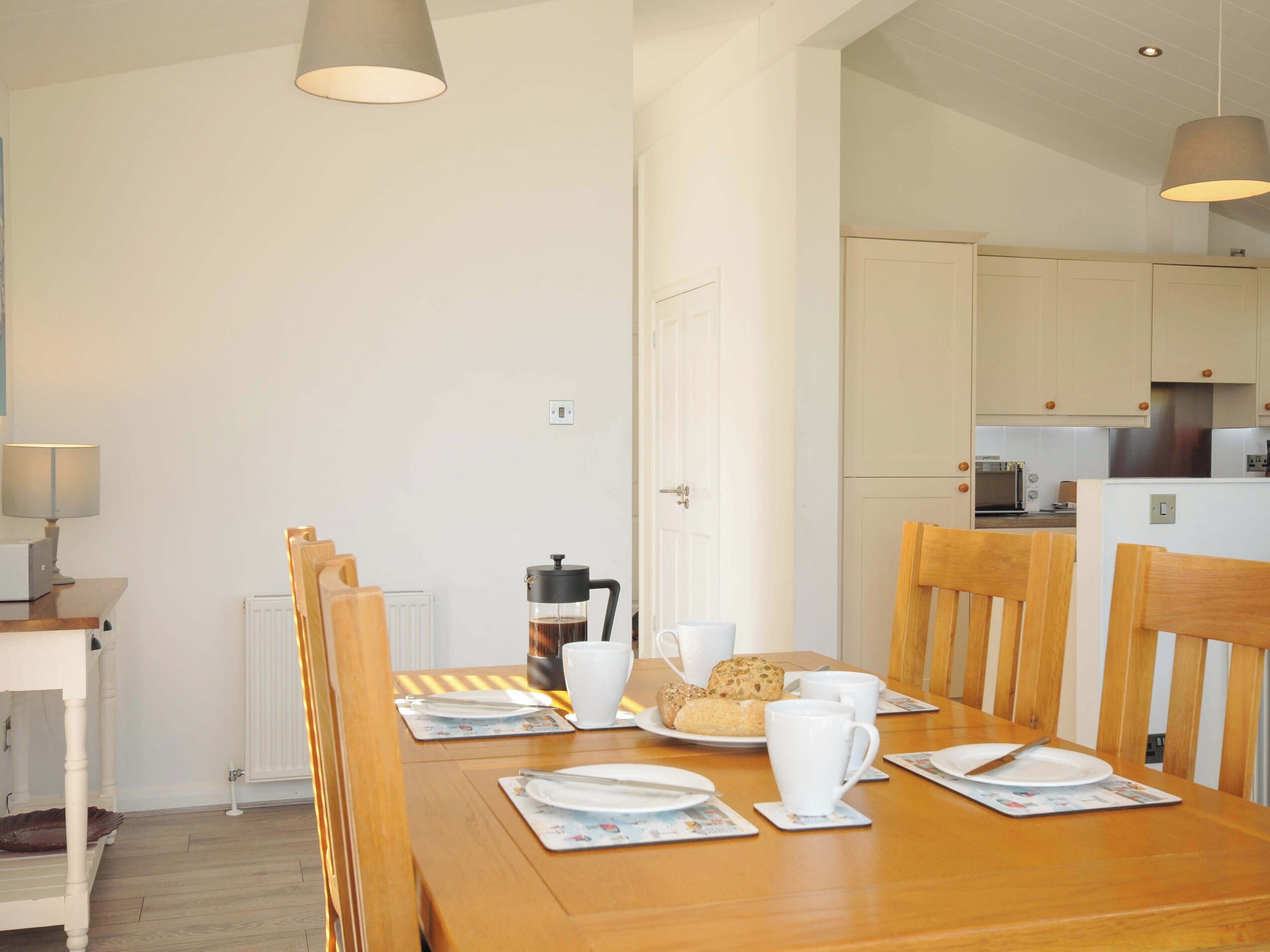 A dining area with a wooden table set for two, a coffee maker, and a plate of cookies.