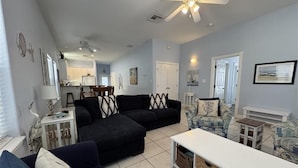 A living room with a black sectional sofa, a white coffee table, and a patterned armchair. There is a ceiling fan, a dining area in the background, and a door leading to another room. - Coastal Cutie by Silver Sands Vacation Rentals (Port Aransas)