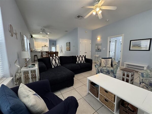 A living room with a black sectional sofa, a white coffee table, and a patterned armchair. There is a ceiling fan, a dining area in the background, and a door leading to another room.