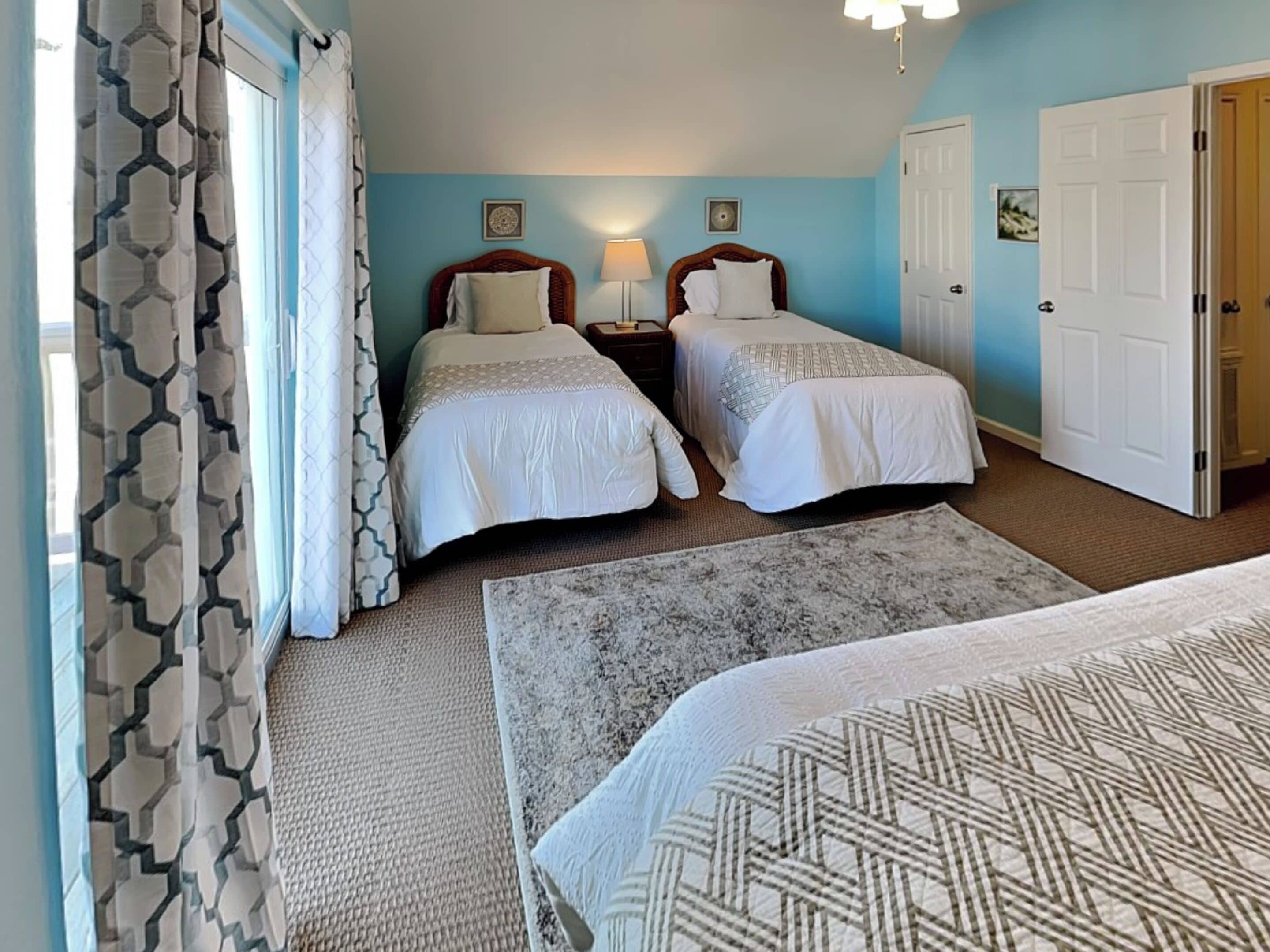 A hotel room with two single beds, a sitting area with a patterned rug, a large window with curtains, and a door.