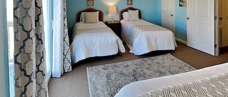 A hotel room with two single beds, a sitting area with a patterned rug, a large window with curtains, and a door.