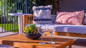 Une table en bois sur laquelle se trouve un bol rempli de plantes et deux verres, un canapé avec des coussins à motifs et, au fond, une clôture en métal.