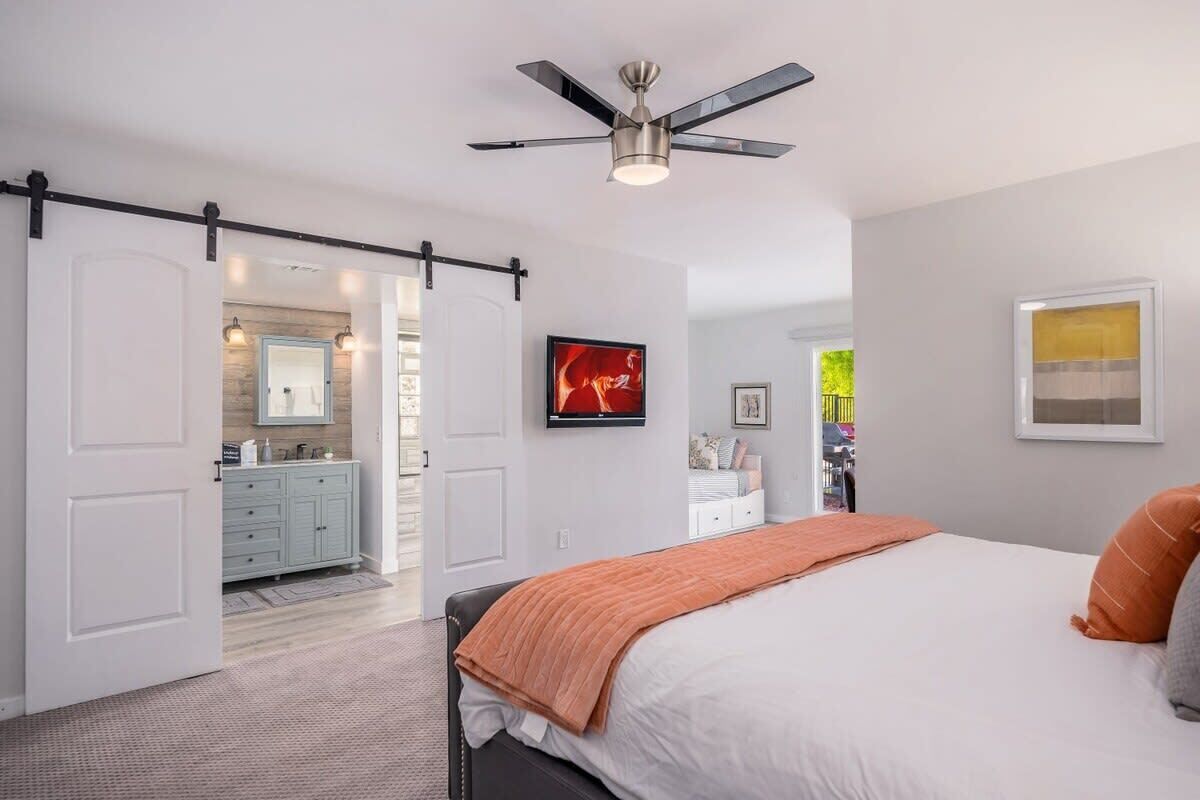 Une chambre à coucher comprenant un lit, un ventilateur de plafond, une télévision et une porte coulissante donnant sur une salle de bain.