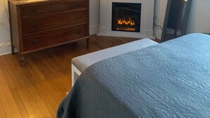 Une chambre avec une commode en bois, un lit recouvert d’une couverture bleue, une cheminée surmontée d’un téléviseur et un tableau accroché au mur.