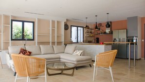 A modern living room with a sectional sofa, two wicker chairs, a glass coffee table, and a kitchen area with a refrigerator, oven, and shelves. - Villa des jacinthes (Tarascon)
