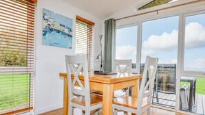 Ein Essbereich mit einem Holztisch und weißen Stühlen, großen Fenstern mit Jalousien und Blick auf eine grüne Landschaft.