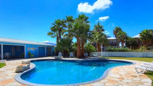 A swimming pool with stone tiles, palm trees, and a blue building in the background.