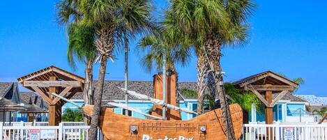 A wooden pirate ship with the name 'Pirates Bay' in front of a building with a blue roof and white fence.